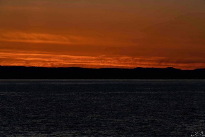 Beautiful sunset photo with shades of rusty orange in the sky above the silhouette of the mountains, with slivery grey sea waters beneath