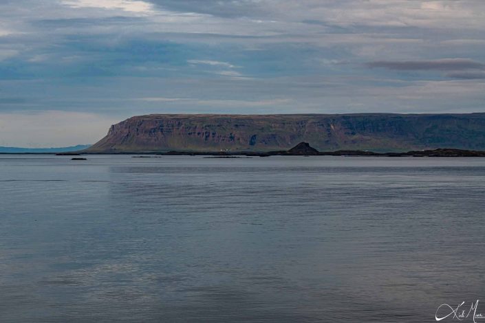 Scenic photo with brown black mountain in the background and silver blue sea water in the front with blue sky above