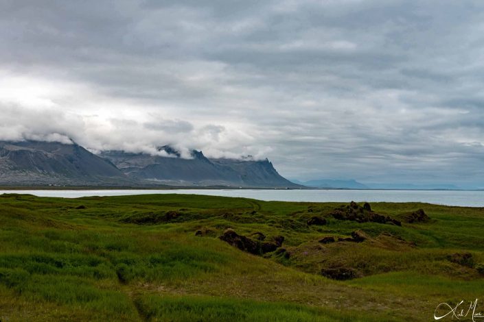 Scenic photo of brown- black mountains with clouds and a green beach by blue waters