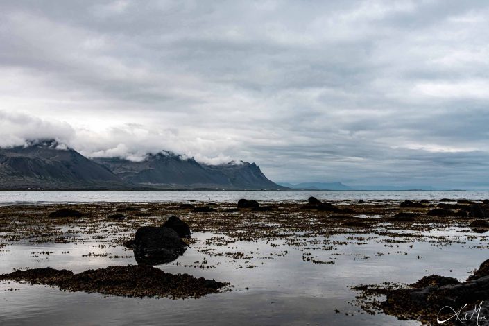 Scenic photo of brown- black mountains with clouds and a beach by blue waters