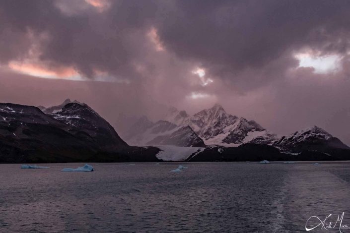 Dramatic photo of what looks like a cloudy and stormy sky over snow capped mountains, and silvery grey sea waters