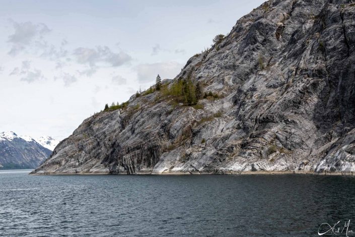 Beautiful photo of a grey-silver mountain with some green trees, along with dark grey-silver waters in the front and snowcapped mountains in the back ground