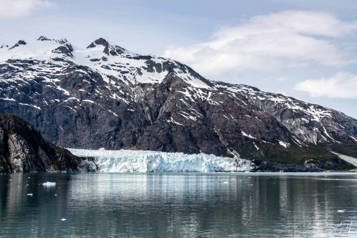 A glacier along with snow capped grey-green mountains with dark grey-silvery waters in the front