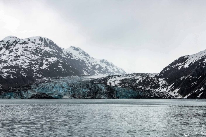 Dark grey-black snow-capped mountains with a glacier and silvery-grey waters in the front