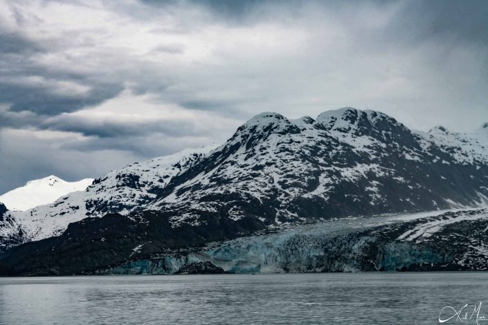 Dark grey-black snow-capped mountains with a glacier and silvery-grey waters in the front