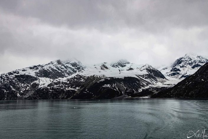 Snow capped dark grey- black mountains with grey- silver waters in the front