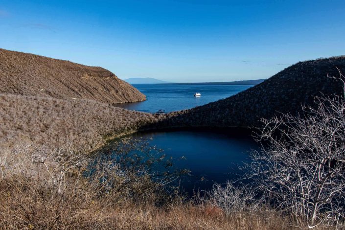 Scenic photo with brown mountains with a blue lake in the middle and deep blue seas and a ship in the background