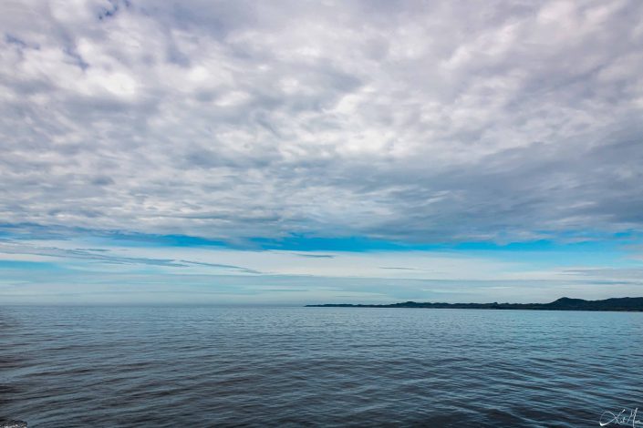 Artistic photo of silvery grey clouds in blue skies above blue silvery waters, with a strip of mountains starting from one side and just ending in the middle